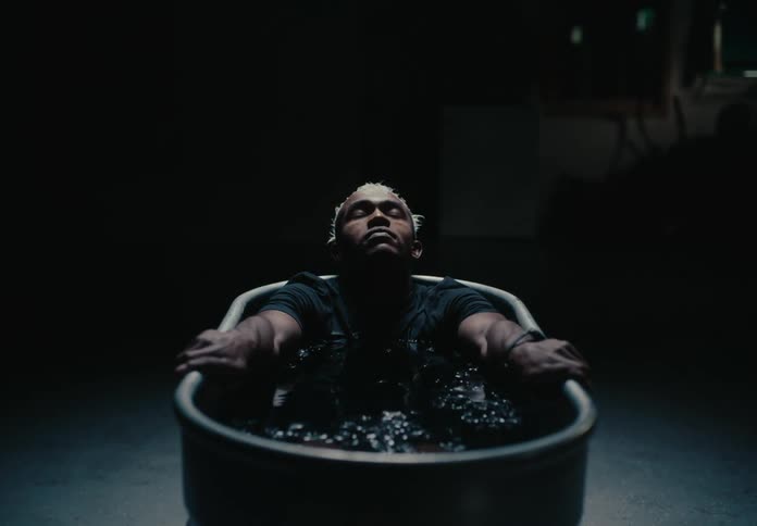 A person sits in a dimly lit tub filled with dark water arms resting on the edges and head tilted back with closed eyes exuding a sense of calm and introspection