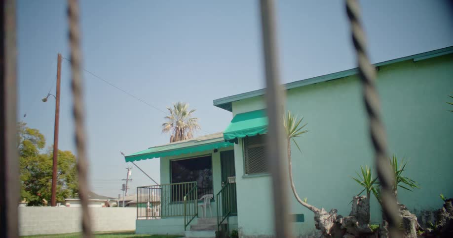 A green house with cloth awnings sits behind a metal fence in a neighborhood