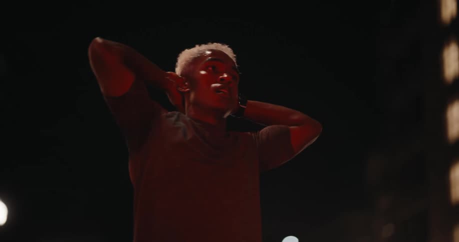 A young man is sweaty and tired and has his hands on the back of his neck as he breathes deeply and it is night time