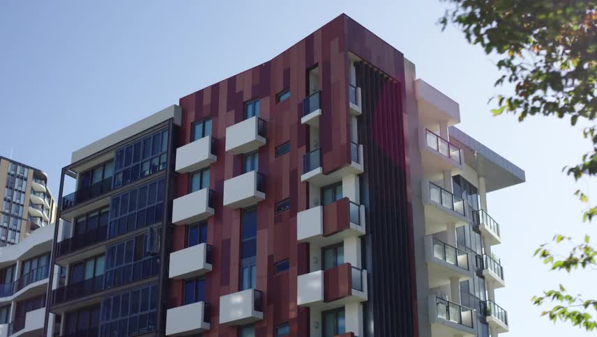A modern multistory apartment building with a mix of brown paneling and white balconies stands under a clear blue sky partially framed by tree branches