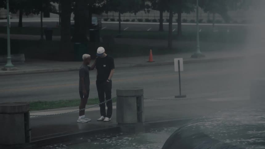 Two men are standing talking next to a fountain that is throwing many drops of water to the sides