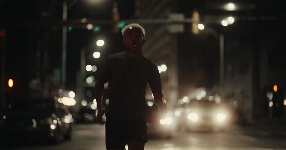 A person running in the middle of a dimly lit urban street at night surrounded by the glow of headlights and streetlights
