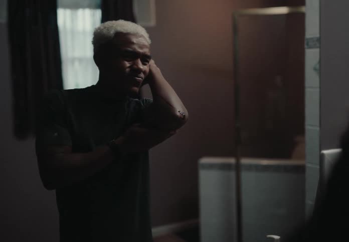 Man stands in a dimly lit room looking into the mirror while touching their arm with a thoughtful or slightly strained expression on their face