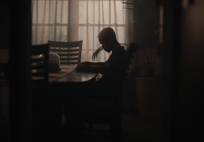 A man is sitting in front of a table and he is thoughtful and darkened due to the low lighting in the room