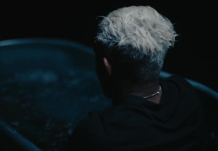 A man with short bleached blonde hair sits in an ice bath his silhouette softened and a delicate necklace glinting subtly
