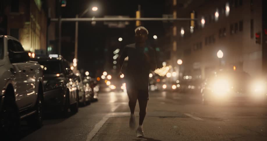 A young man is running on a paved street at night with several cars parked on the left and some walking on the right