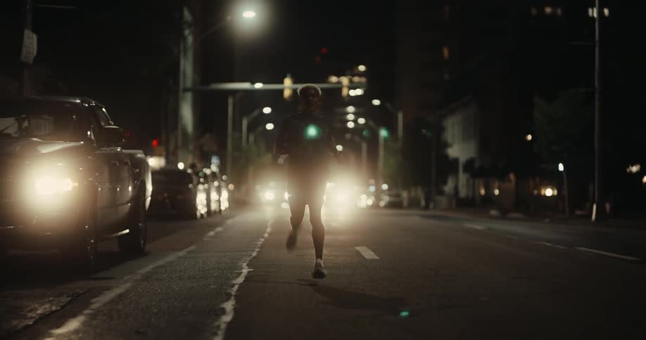 Man is running down a city street at night illuminated by the bright headlights of an approaching car with streetlights and buildings casting a dramatic urban backdrop