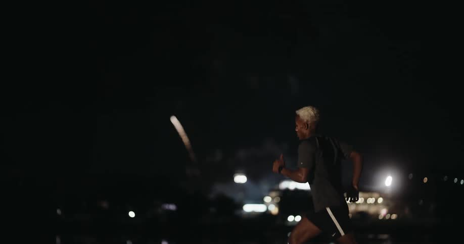 A young man is running quickly one night and behind him there are fireworks being exploded in the sky