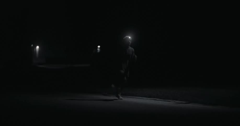 A man running in the dark illuminated by a headlamp with faint streetlights visible in the background