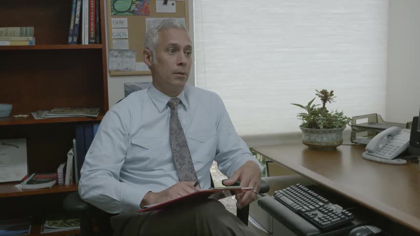 Man sitting in an office holds a clipboard and talks