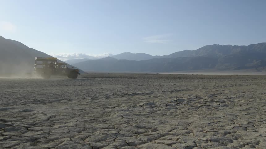 A vehicle drives across a large dirt field with mountains in the distance
