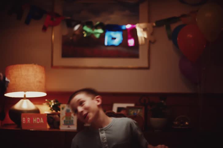 A young boy seated at a table in a warmly lit room smiling joyfully and moving his head while being filmed during a birthday celebration with colorful decorations and cards in the background