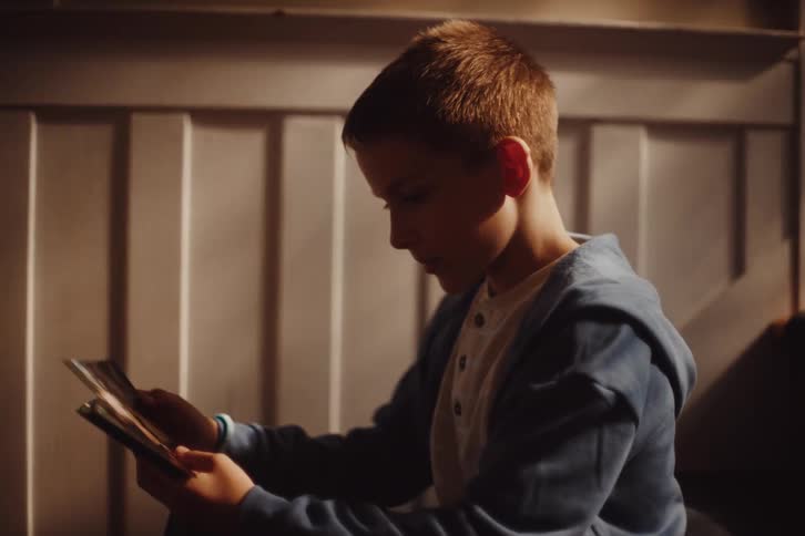 A young boy sitting in a softly lit room intently focused on photographs he is holding in his hands with his profile illuminated against a plain paneled background
