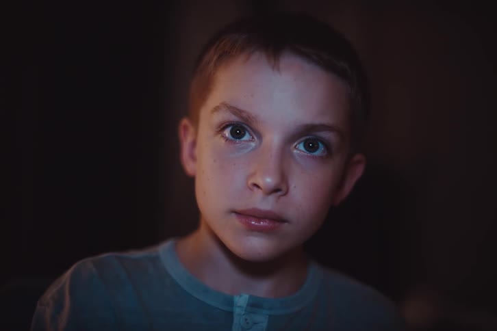 A boy wearing a gray shirt watches a screen with a wide smile