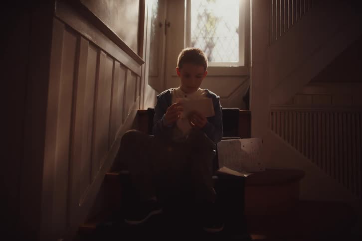 A boy sits on the stairs reading a letter and then looking at photographs