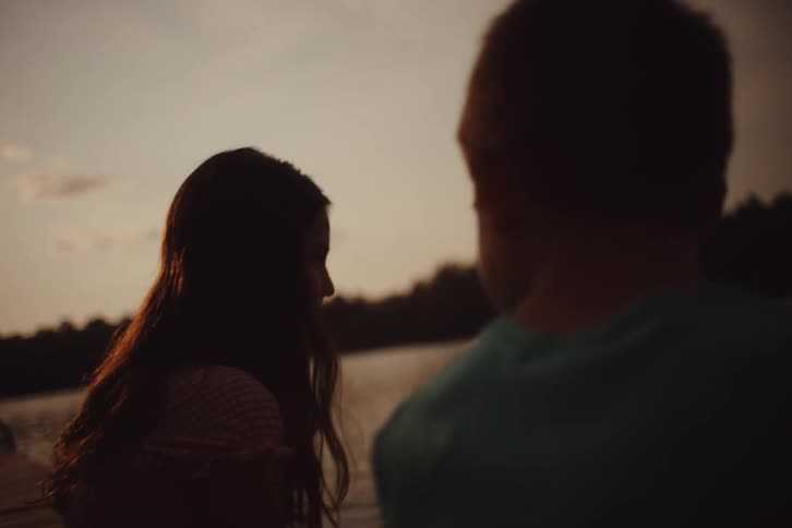 A child wearing a blue shirt and a woman with wavy hair sits by the lake at dusk