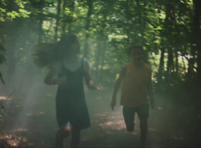 Two children run through a hazy forest their clothing and hair slightly blurred by the movement and fog