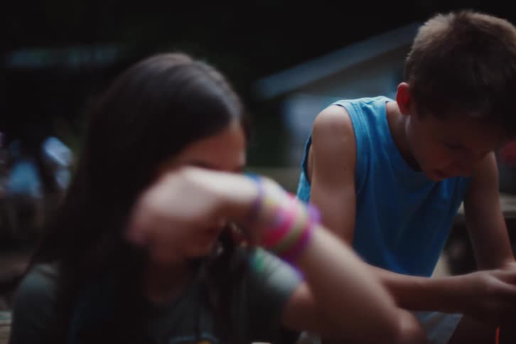 Two children are sitting outdoors at night at a campsite focused on crafting bracelets