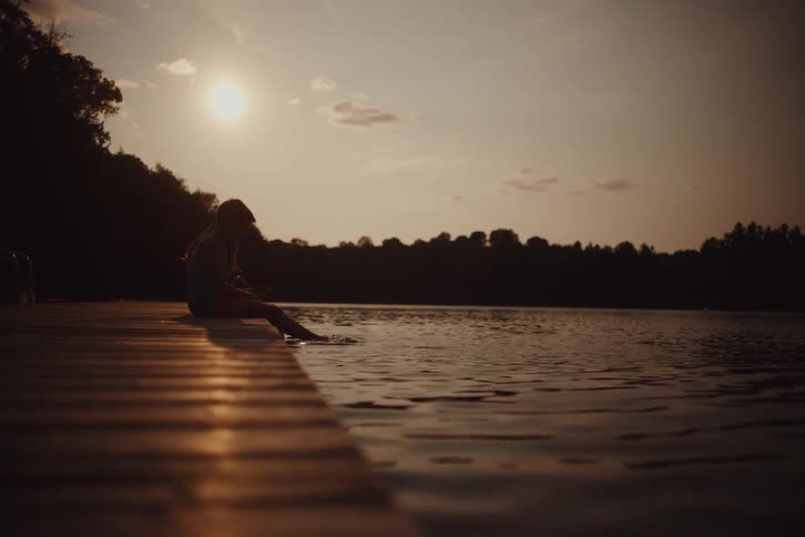 Two children sitting on a pier dipping their feet in a lake in the late afternoon