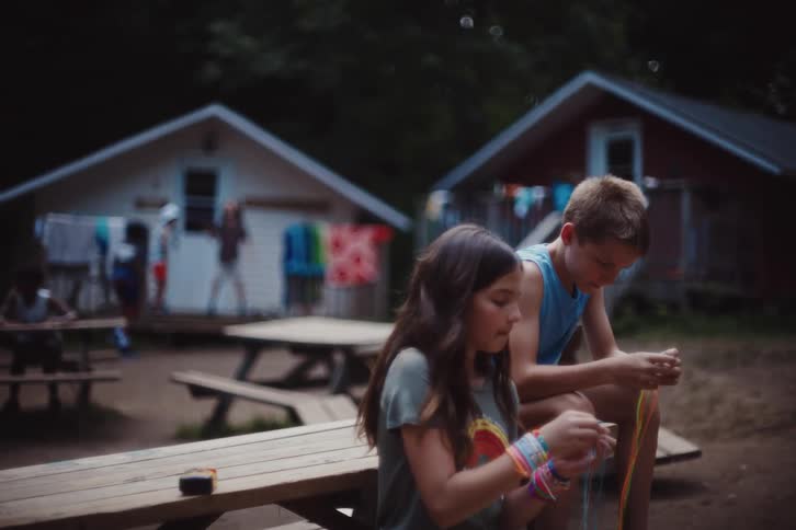 A girl and a boy sit on a picnic table doing craftwork as people goof around in the background