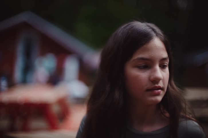 A young girl with long dark hair gazes downward thoughtfully set against the blurred backdrop of a wooden cabin in a natural outdoor setting