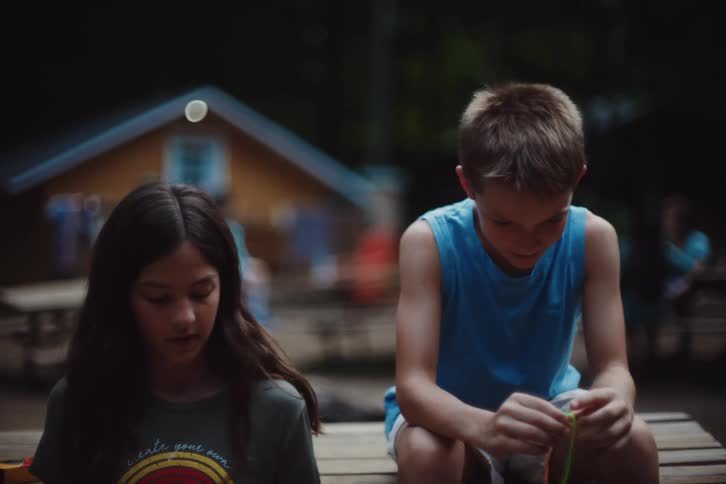 Two children a girl and a boy sit outdoors at dusk at a campsite working on crafting bracelets