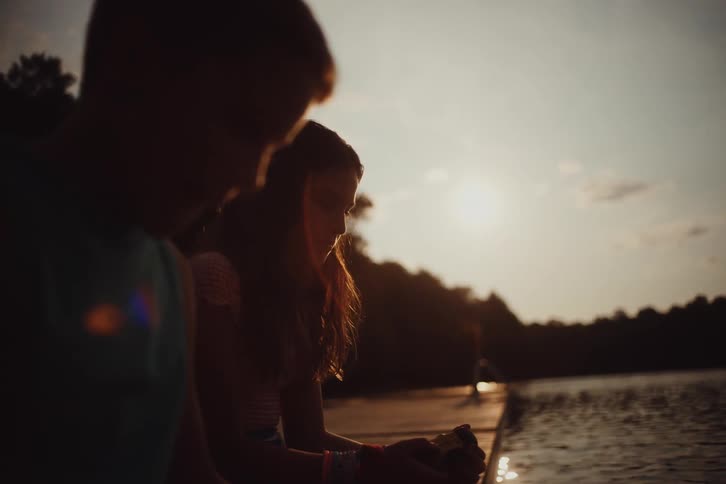 A little boy and a little girl next to her and they are both sitting on a pier at the edge of a lake and they are looking down