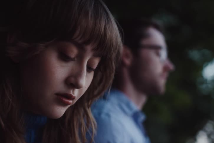 A young woman looks down pensively while a man stands in the blurred background