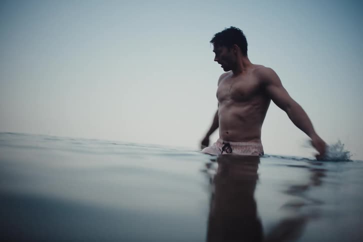 A shirtless man is standing in waistdeep water and moving his hands on the surface of the water