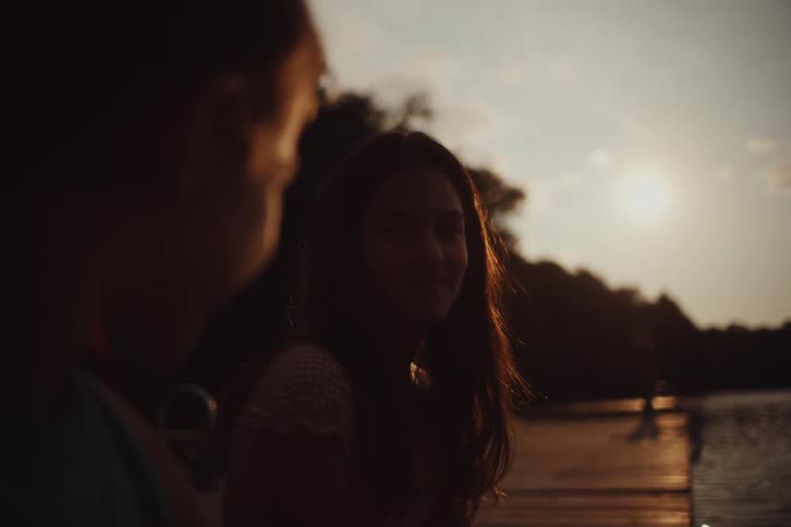 Silhouetted figures of people sit on a lakeside dock as the sun sets with a focus on a teenage girl
