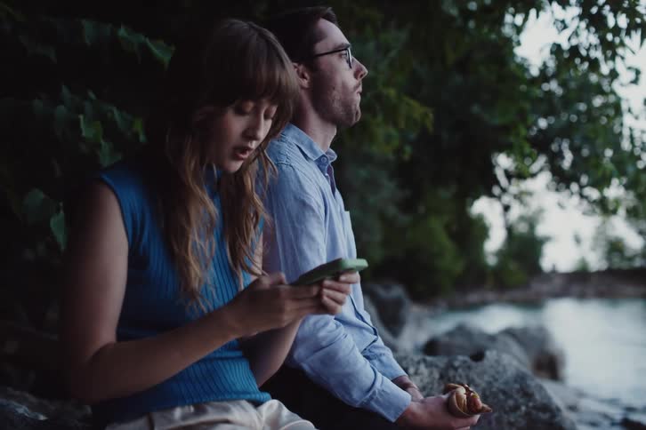 A woman uses her cell phone and a man eats by the stream