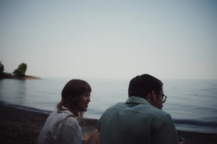 A man and a woman are sitting on the sand in front of a beach and are chatting and having fun while the man is trying to eat something