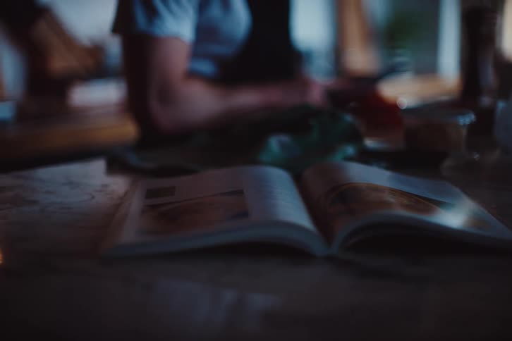 An instruction book is open on a table and there is a person in the background who is fiddling with something on the table