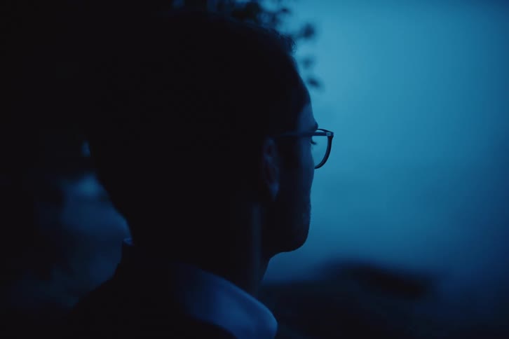 A man wearing glasses gazes out at a bluish toned twilight landscape