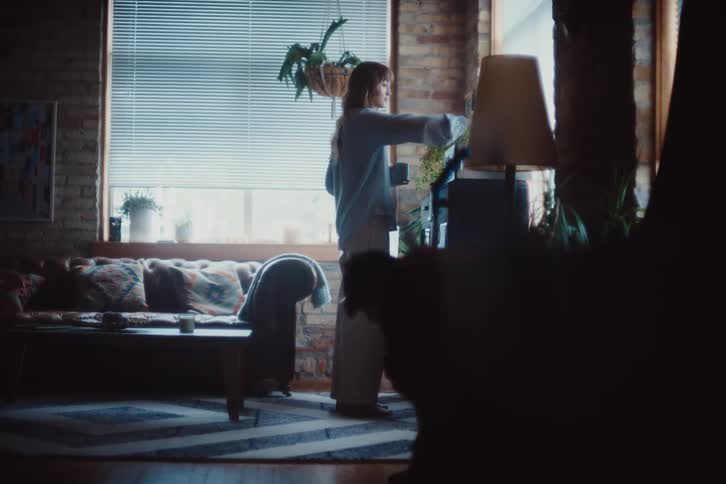 A cozy living room with exposed brick walls a tufted sofa with patterned cushions and a woman standing near a window adjusting plants in a softly lit serene atmosphere