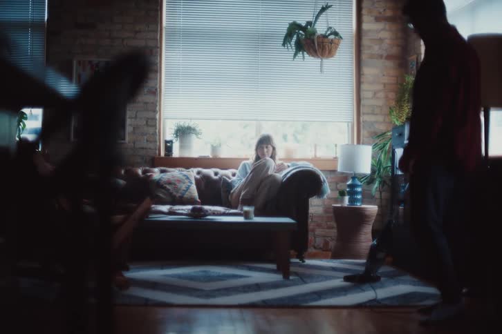 A person standing using a vacuum cleaner to clean a carpet while another person sits on a sofa