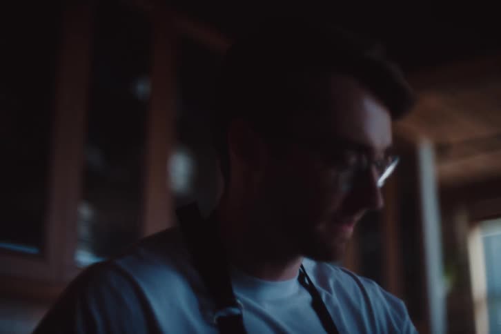A young man wearing reading glasses and a black apron looking down while adjusting something with his hands