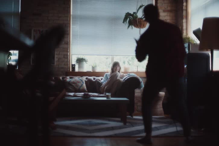 A person is standing cleaning a carpet and starts dancing for the person sitting on the sofa