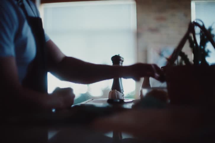 A man in an apron standing and picking up some food ingredients from a cutting board on the table