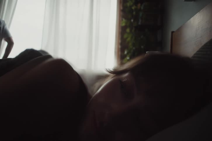 A man opens the curtains as the woman rests in bed