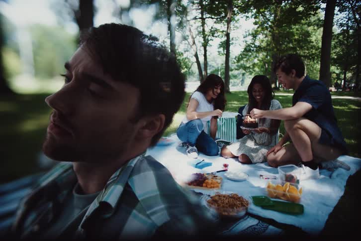 A young man sits apart from his friends looking sad and lonely while his friends laugh while lighting a birthday cake on a blanket filled with food dishes