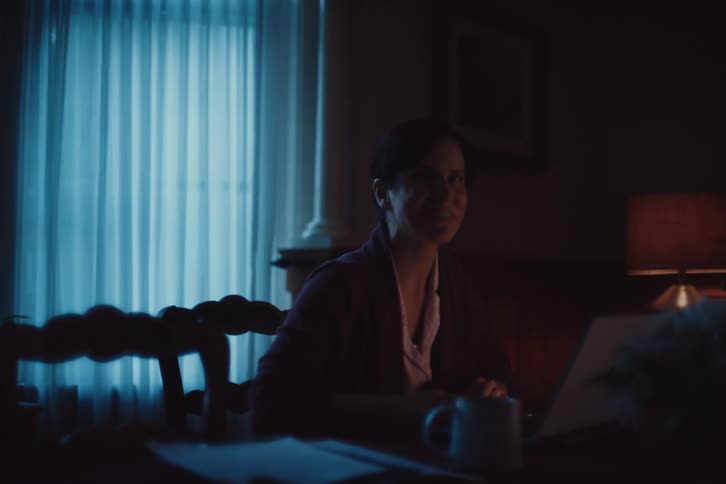 A woman in a purple robe sits at a table working on a laptop in a dimly lit room at night