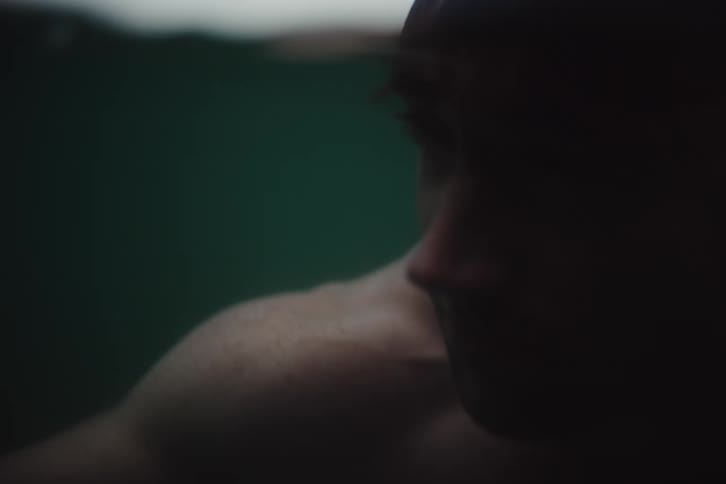 A muscular man diving underwater with his upper body partially visible in a blurry cinematic perspective