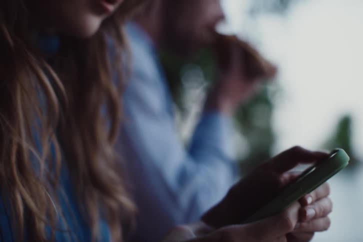 A woman is texting looking intently at her phone while a man in the background eats and appears distracted
