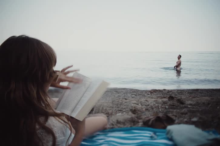 Woman lying on the sand reading a book and a man in the background at the sea on the beach