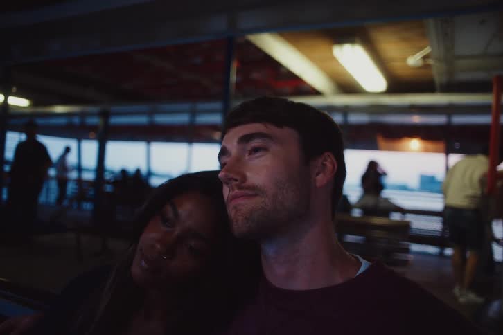 A tender moment between two people with a woman resting her head on a mans shoulder in a softly lit indoor setting with blurred figures and windows in the background