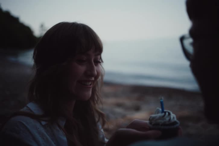 A woman smiling and receiving a cupcake with a lit candle is enjoying a special moment with a man on a beach at twilight