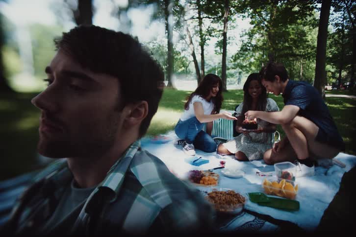 Three people are having a happy picnic and on the left side there is a mans face who has his back to them and he is sad