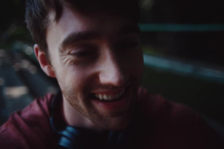 The face of a young man who is talking to someone in front of him and he gives small smiles while he talks and there is a headphone around his neck