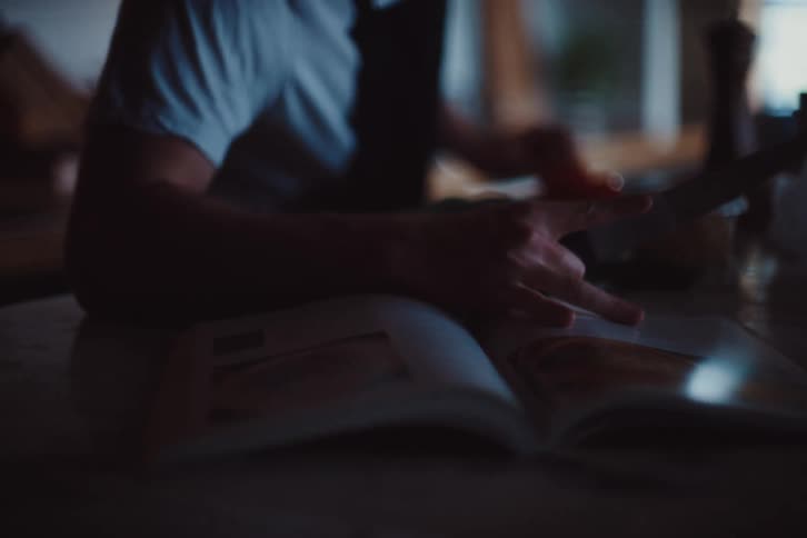 Person holding a knife while cooking and reading a recipe book next to it